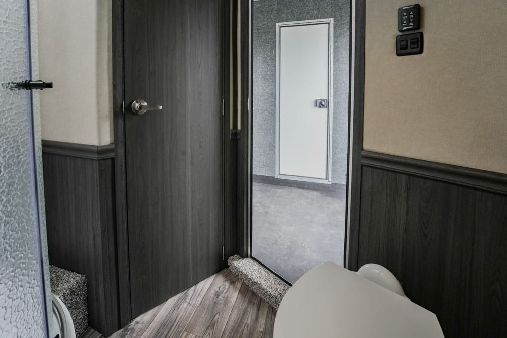 Interior bathroom with dark wood paneling, a closed toilet seat in the foreground, and a doorway to another room in the back.
