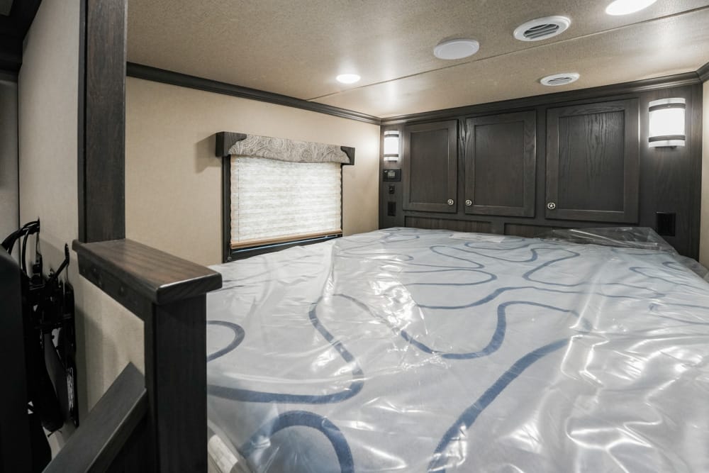 RV bedroom with a large bed covered in plastic, dark wood cabinets on the back wall, and soft ceiling lights.