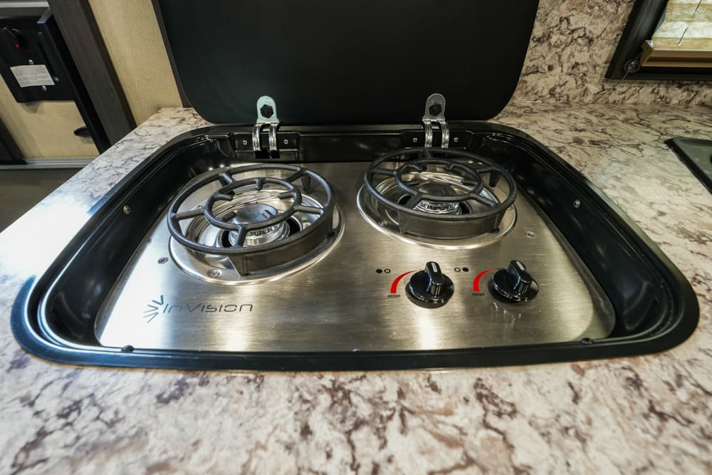 Two-burner gas stove in an RV with a hinged lid, stainless steel surface, and marbled countertop.