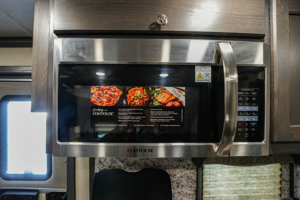 Stainless steel microwave with a black door and control panel on the right, labeled Contoure, mounted in a wood cabinet above a counter.