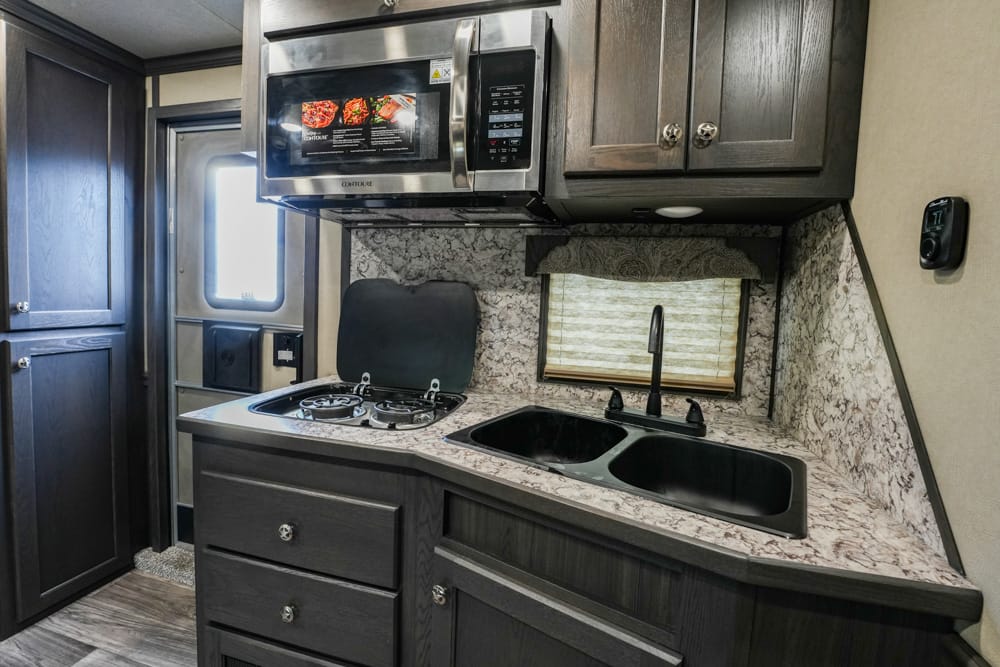 Compact RV kitchen with a two-burner stove, microwave above, dark cabinets, and a double sink under a small window.
