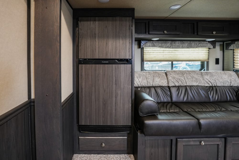 Interior of an RV living area with a tall wood-front refrigerator, dark cabinets, and a black leather sofa by a window with blinds.
