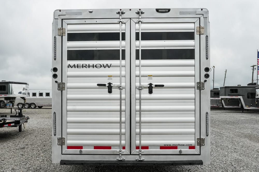 Rear view of a silver Merhow horse trailer with double doors and latches, parked on a gravel lot.