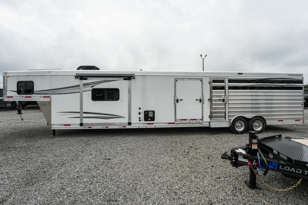White horse trailer with gray decorative graphics parked on a gravel lot; side access door and small window along the midsection, chrome ventilated stalls at the rear, and a front gooseneck area visible with a trailer hitch in the foreground.