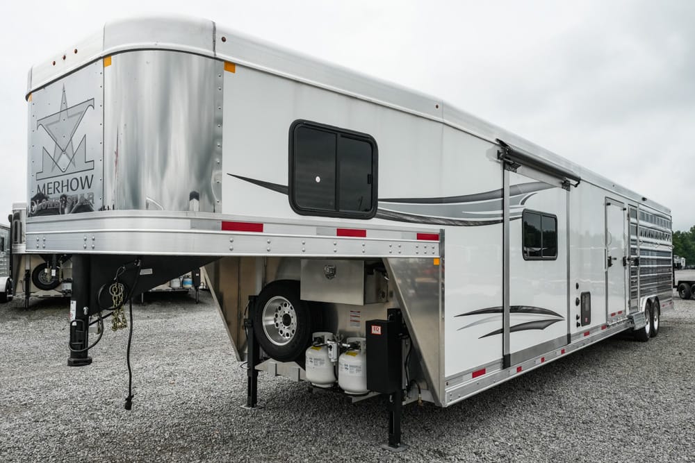Gooseneck horse trailer with living quarters by Merhow, parked on gravel; shows hitch and propane tanks under the trailer.
