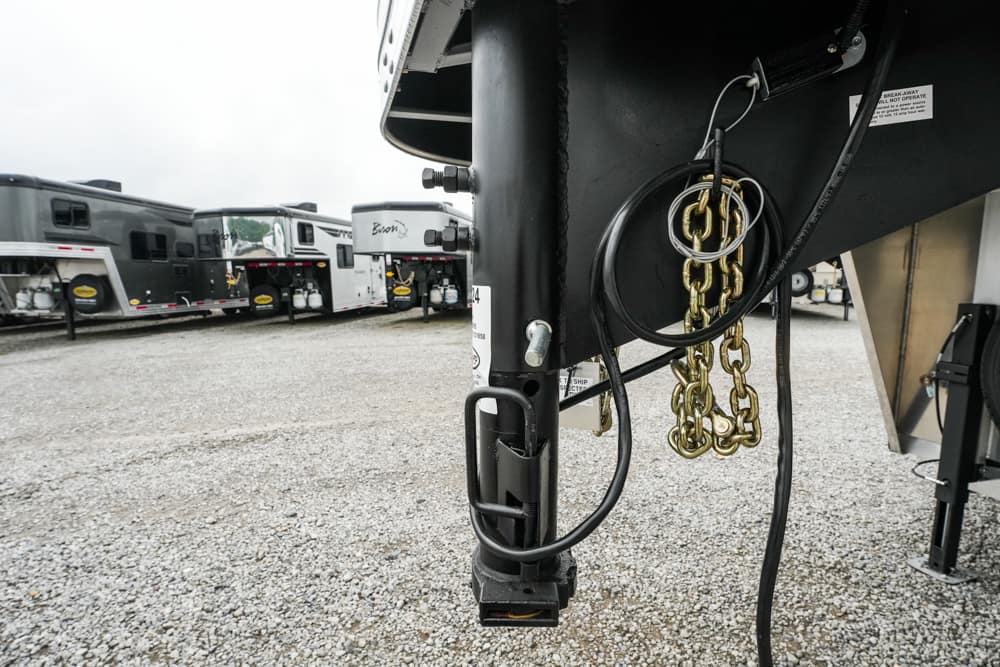 Close-up of a gooseneck trailer hitch with safety chains and electrical cables, parked among RVs on a gravel lot.