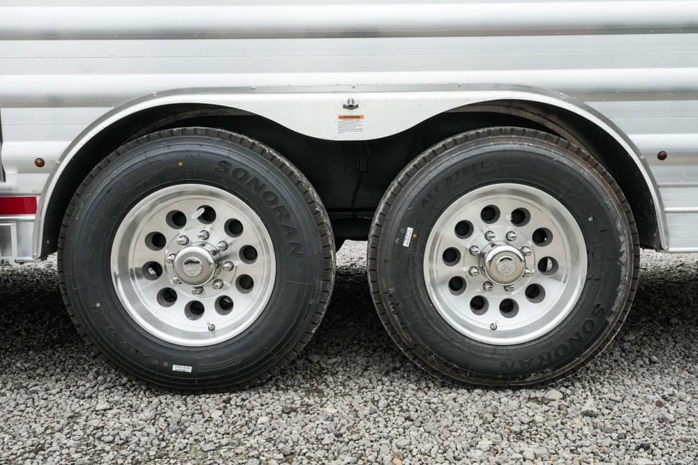 Tandem axle trailer wheels: two silver rims with lug nuts on a gravel surface.