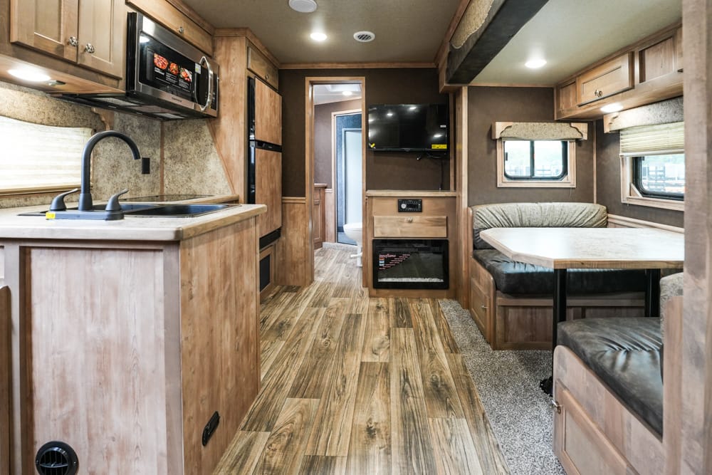 Interior of a modern RV: kitchen on the left with a sink, microwave, and wooden cabinets; dining booth on the right with a table.