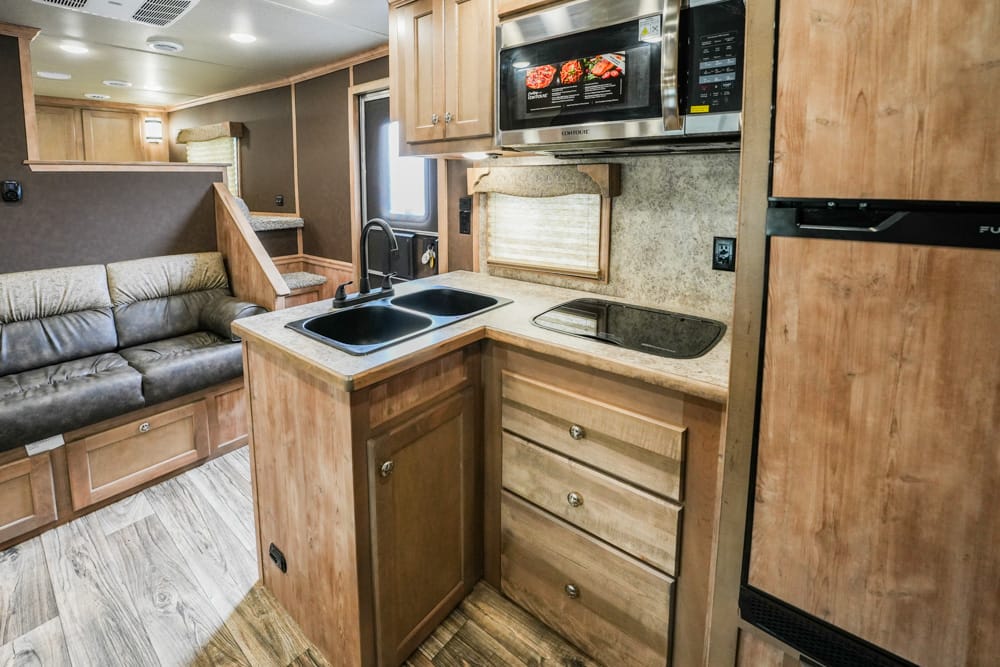 Interior of a motorhome kitchen: double sink, black glass stovetop, microwave above, and light wood cabinets with a beige sofa in the living area nearby