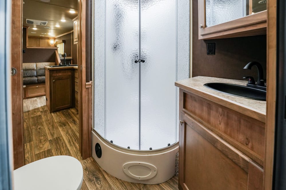 Interior of an RV bathroom showing a curved frosted shower enclosure, wood cabinetry, and a sink on a beige countertop; toilet in foreground.