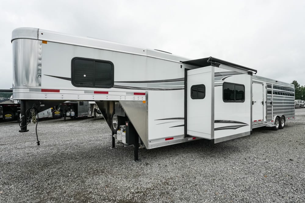 White gooseneck living-quarters trailer with a side slide-out, parked on a gravel lot, two axles at the rear and multiple windows in view