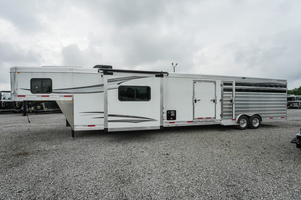 Long white enclosed horse trailer with living-quarters, side windows, and multiple access doors parked on a gravel lot under a cloudy sky.