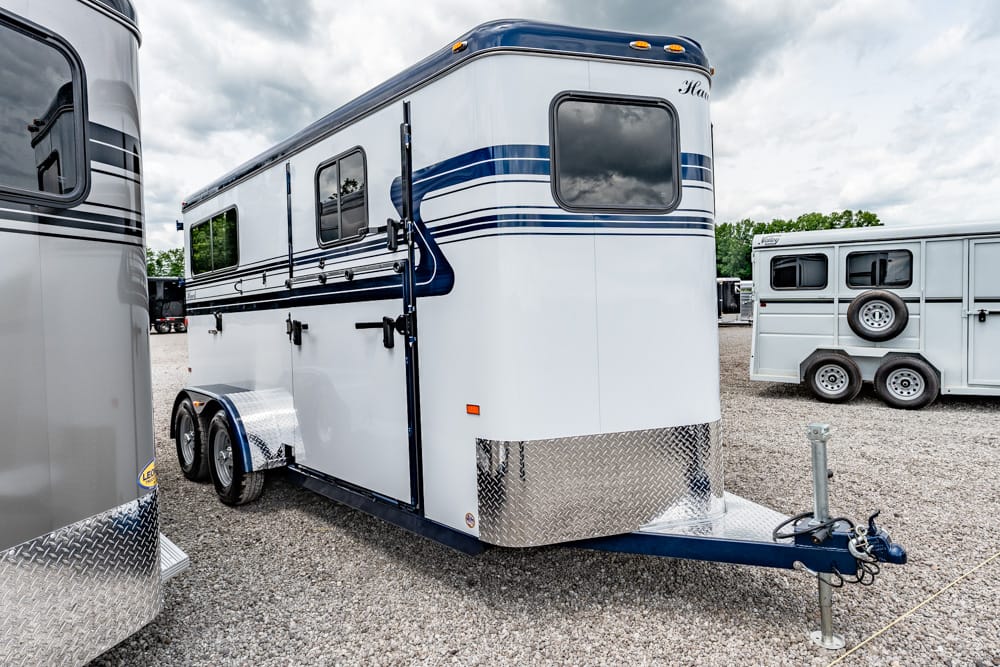 White horse trailer with blue stripes and a diamond-plate front, parked on a gravel lot with other trailers nearby.