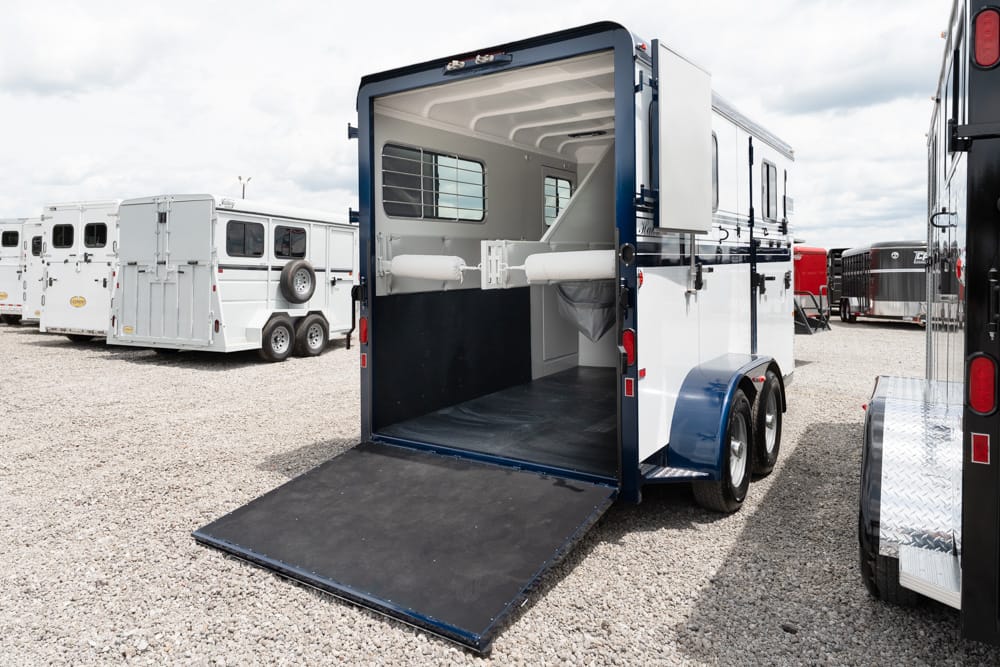 Rear ramp horse trailer with blue trim, showing empty interior stalls and a divider, parked in a gravel lot among other trailers