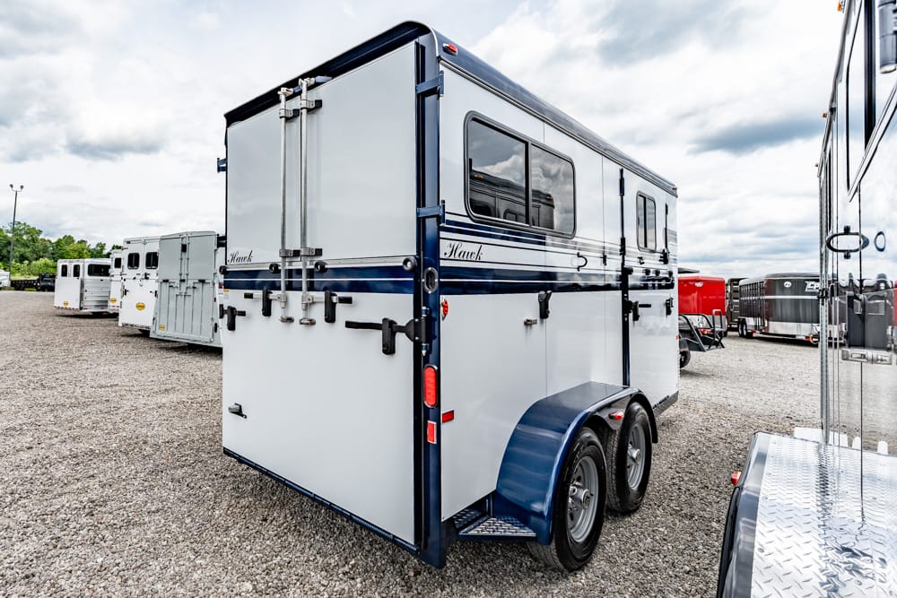 Rear view of a white horse trailer with blue trim and dual axles parked on a gravel lot, with other trailers in the background