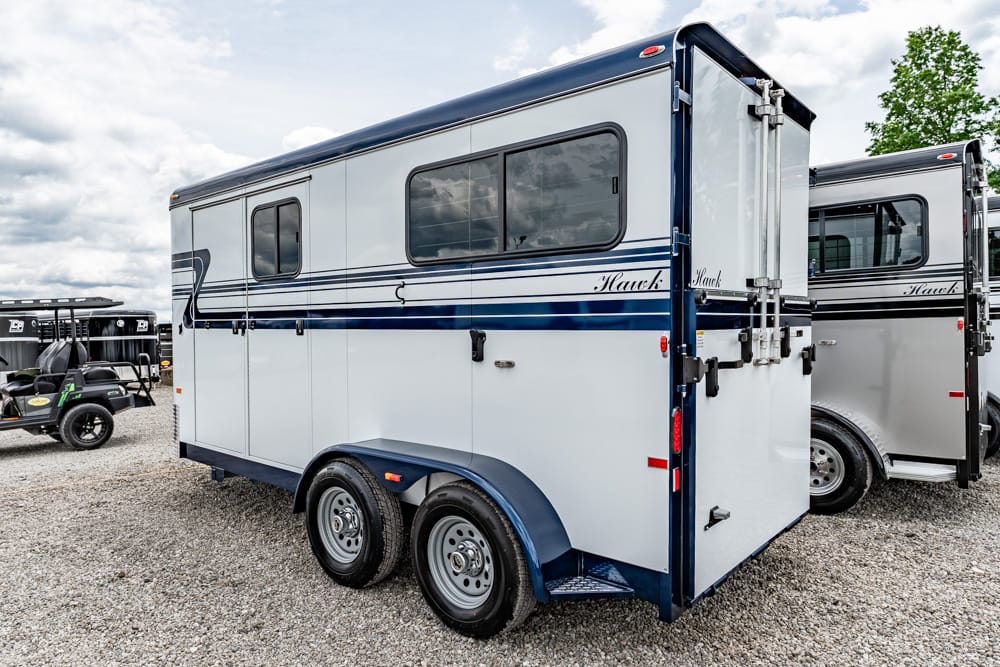 White and blue 2-axle horse trailer on a gravel lot, with another trailer beside it and a cloudy sky above.