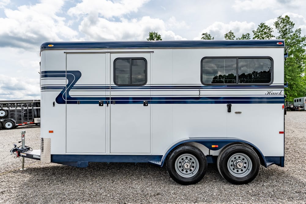 White horse trailer with blue stripes and dual axles parked on a gravel lot, hitch visible at the front.