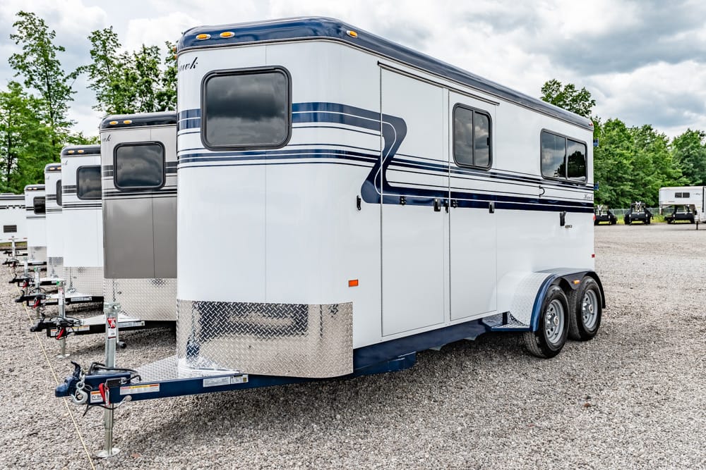White horse trailer with blue stripes and a diamond-plate front, parked on a gravel lot with trees in the background a trailer dealership setting.