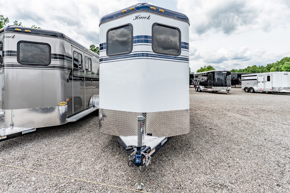 White horse trailer with blue stripes on a gravel lot, front diamond-plate hitch area and tow bar, other trailers in the background under a cloudy sky.