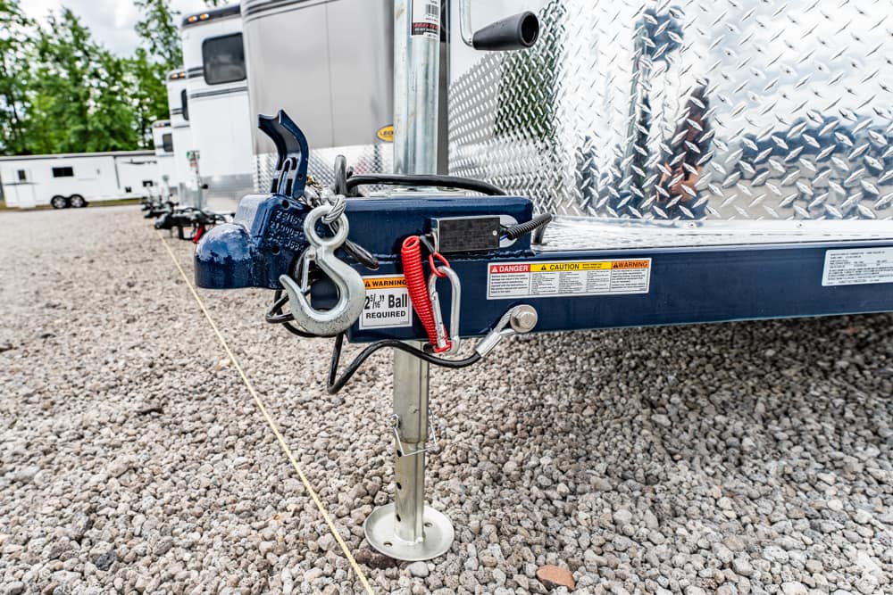 Trailer hitch with safety chains and coupler on a diamond-plate trailer, parked on a gravel lot with other trailers in the background.