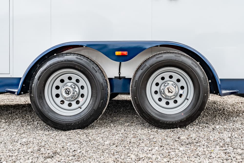 Double trailer wheels with silver rims under a blue and white trailer on gravel surface (tandem axle)