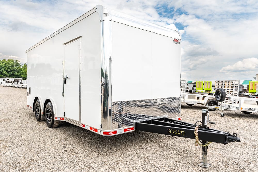 White enclosed cargo trailer with tandem wheels and a hitch, parked on a gravel lot.