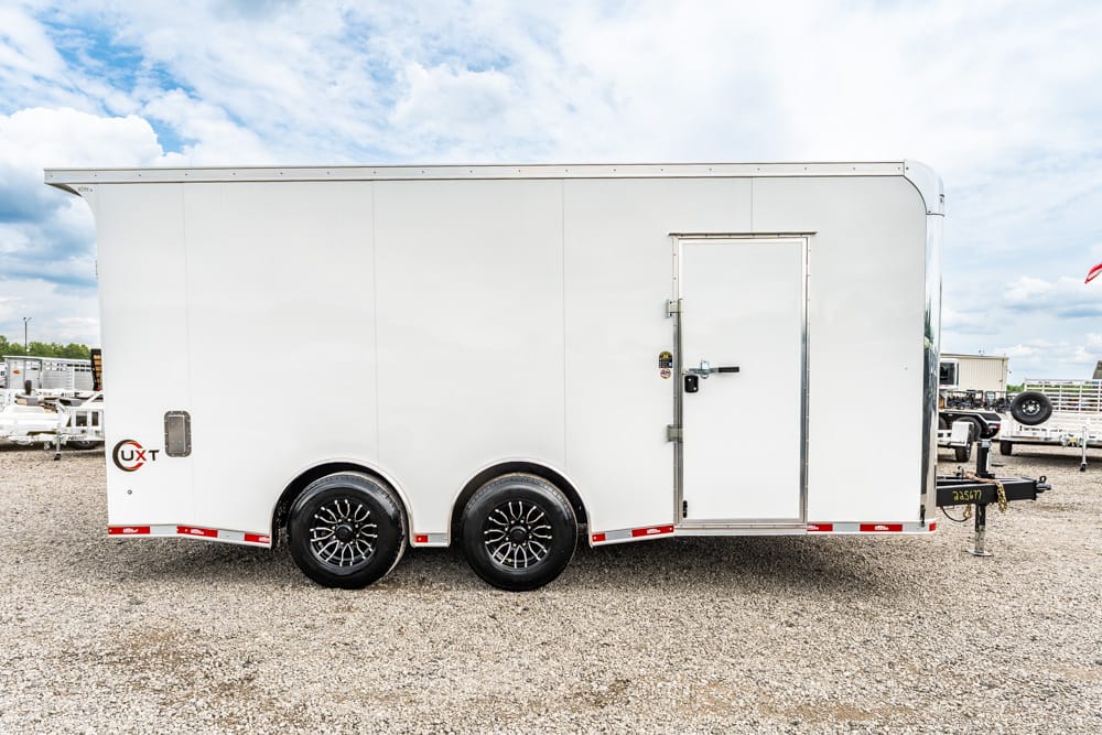 White enclosed utility trailer with two axles parked on a gravel lot; side door closed and hitch visible.