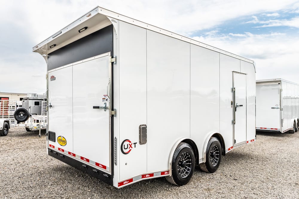 White enclosed cargo trailer with dual rear doors and black alloy wheels on a gravel lot. An outdoor vehicle yard is in the background.
