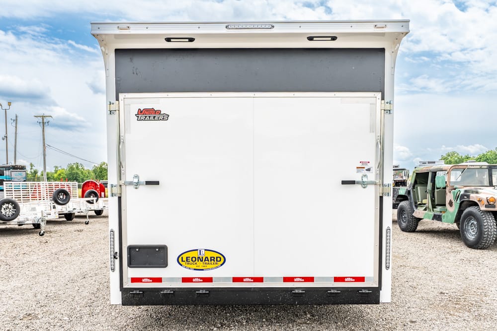 Rear view of a white enclosed trailer with double doors, featuring a Leonard trailer decal and red reflective striping on a gravel lot.