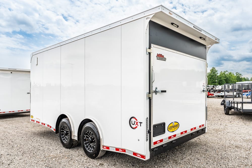 White two-axle enclosed cargo trailer with dual rear doors and red reflective trim on a gravel lot, under a cloudy sky.