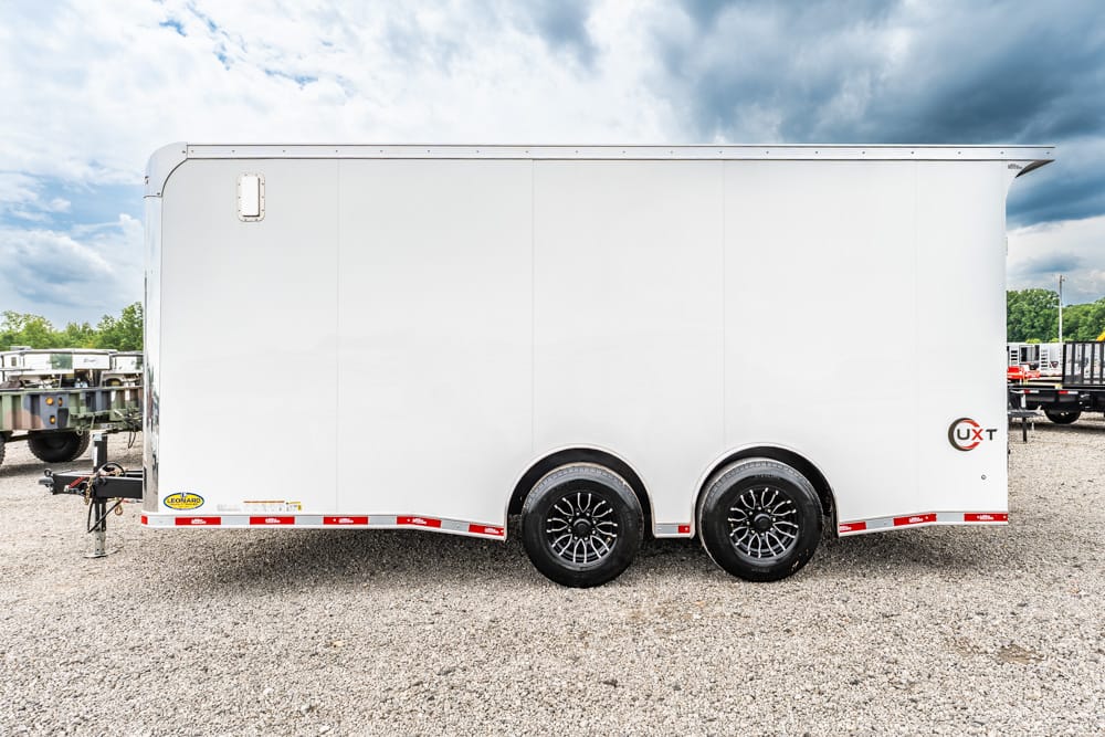 White enclosed cargo trailer with dual axles and black spoke wheels on a gravel lot under a cloudy sky