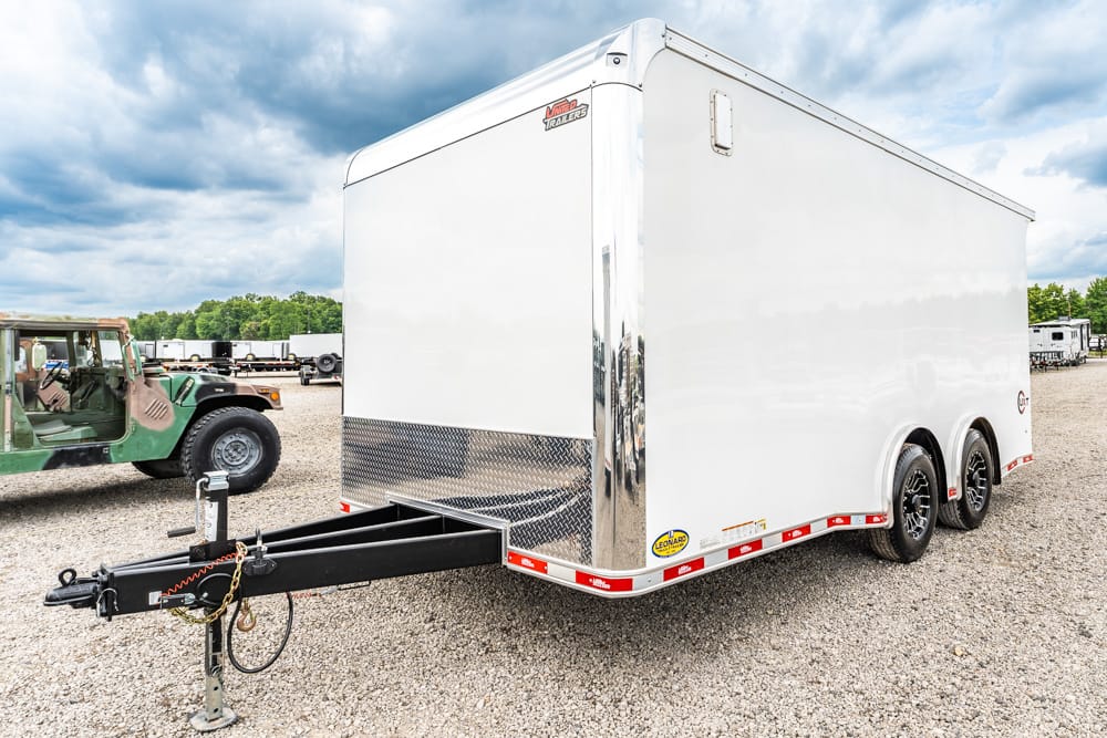White enclosed cargo trailer with dual axles and chrome front, parked on a gravel lot; visible tow hitch, safety chains, and chains.
