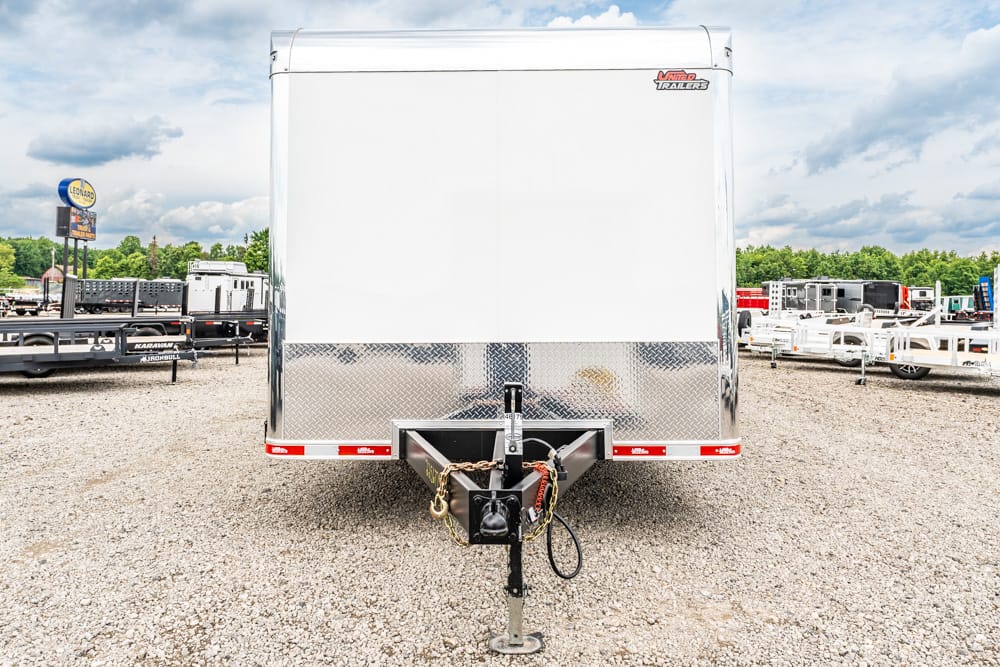 Rear view of a large white enclosed cargo trailer with diamond-plate lower front, parked on a gravel lot with other trailers in the background, and a trailer hitch at the bottom.