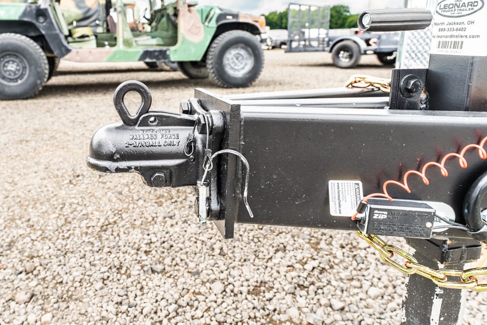 Close-up of a trailer hitch with the coupler and safety chains on a black metal frame; gravel lot and trucks in the background.