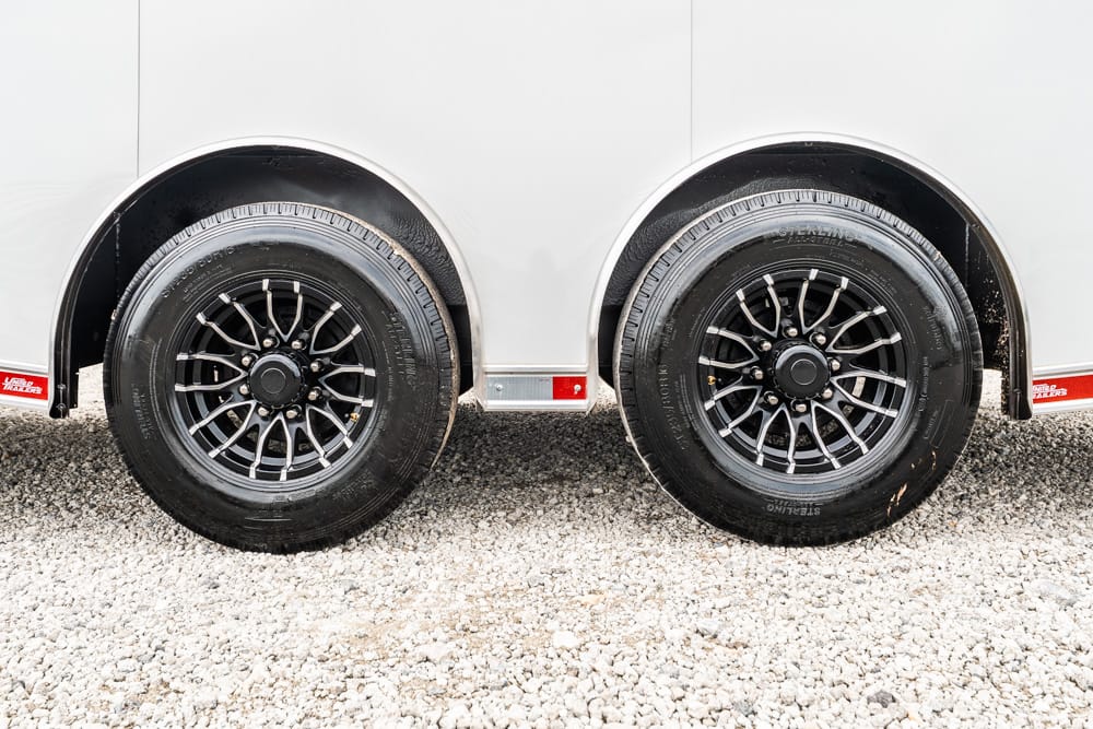 Two black alloy trailer wheels on a silver trailer, parked on gravel ground.
