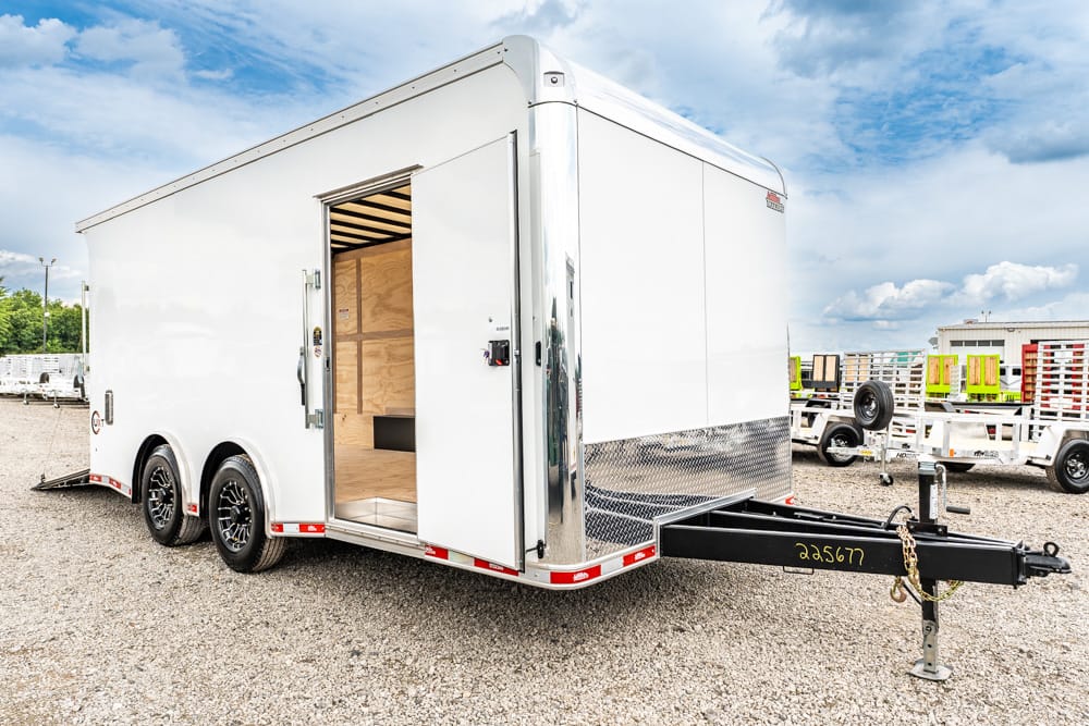 White enclosed cargo trailer with open side door, parked on a gravel lot; tandem axles and front hitch visible in foreground.