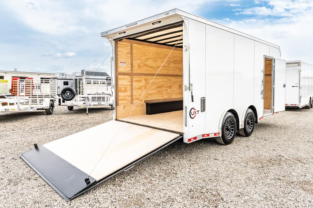 White enclosed cargo trailer with its ramp lowered, parked on a gravel lot; plywood interior visible inside the trailer.