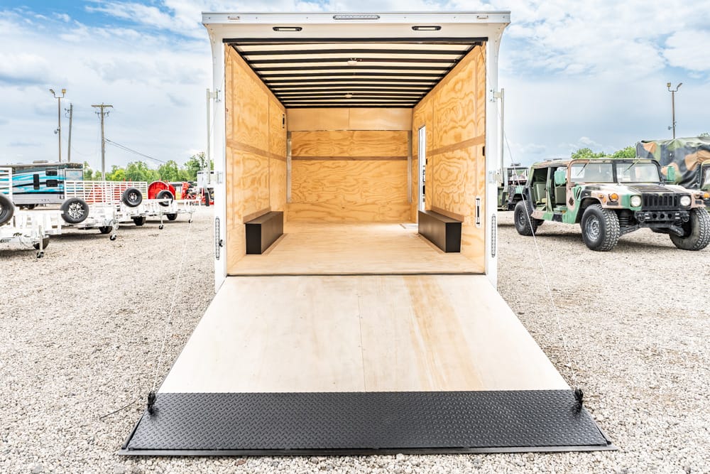 Open empty cargo trailer with plywood interior and a lowered ramp on a gravel lot, with vehicles in the background.