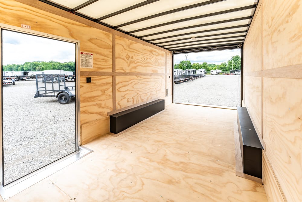 Interior of a plywood-walled trailer with long black benches along the sides, open rear door showing a gravel lot with trailers outside.