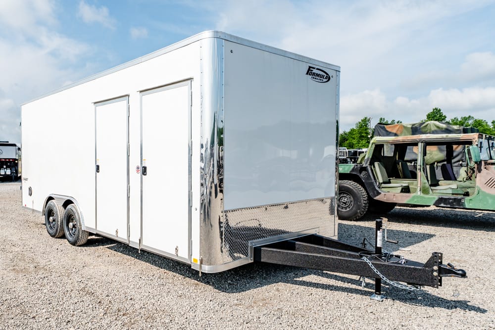 White enclosed dual-axle trailer on a gravel lot, attached hitch and front A-frame tongue visible; military vehicle nearby.