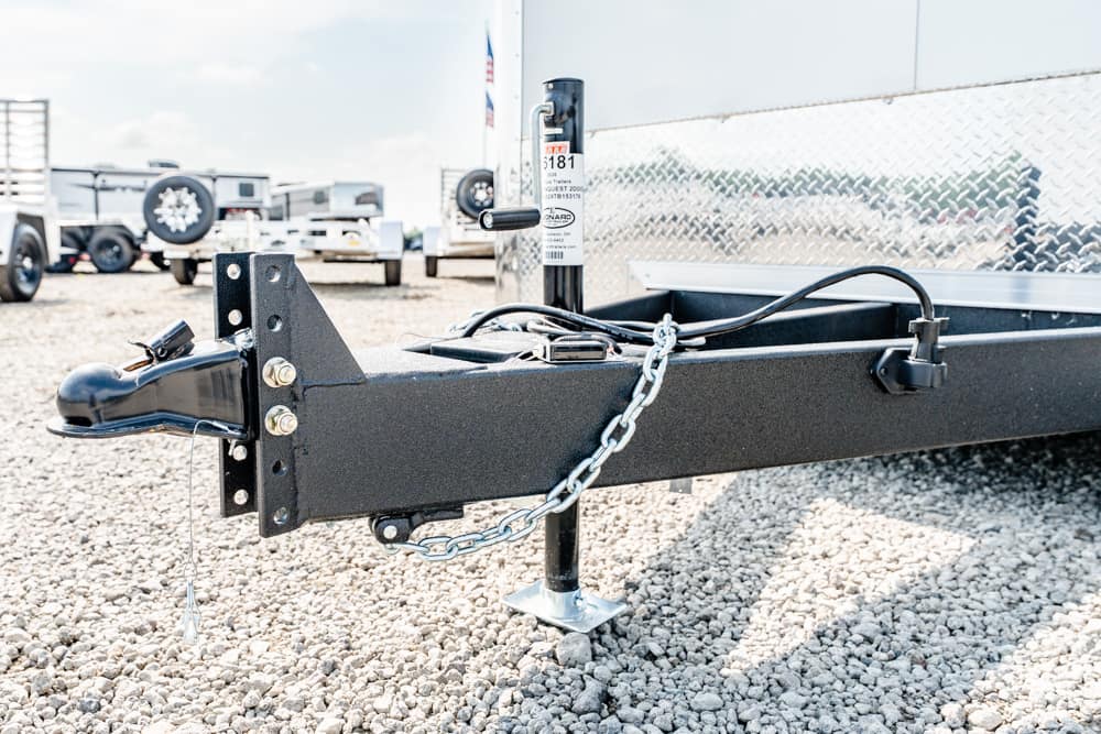 Close-up of a trailer hitch with safety chain and jack on a black trailer frame, resting on gravel in a trailer yard.