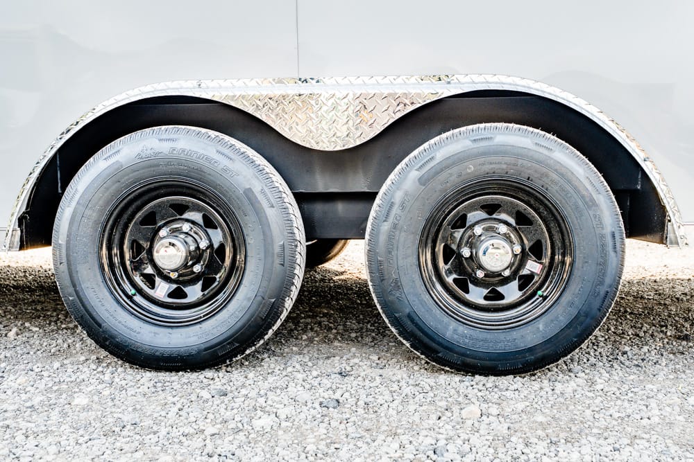 Dual trailer wheels with black rims and a diamond-plate fender on a gravel surface