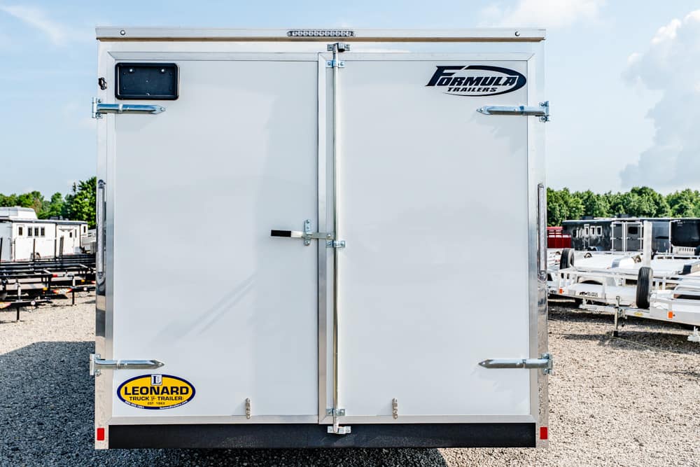 Rear view of a white enclosed cargo trailer with double doors and metal latches, brand logos on top right and bottom left, in a trailer yard.
