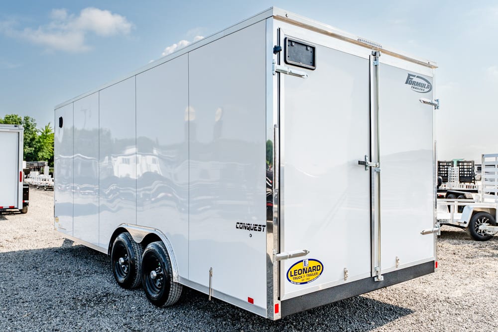 White enclosed cargo trailer with dual rear doors on a gravel lot, chrome hardware and brand decals visible at the back.