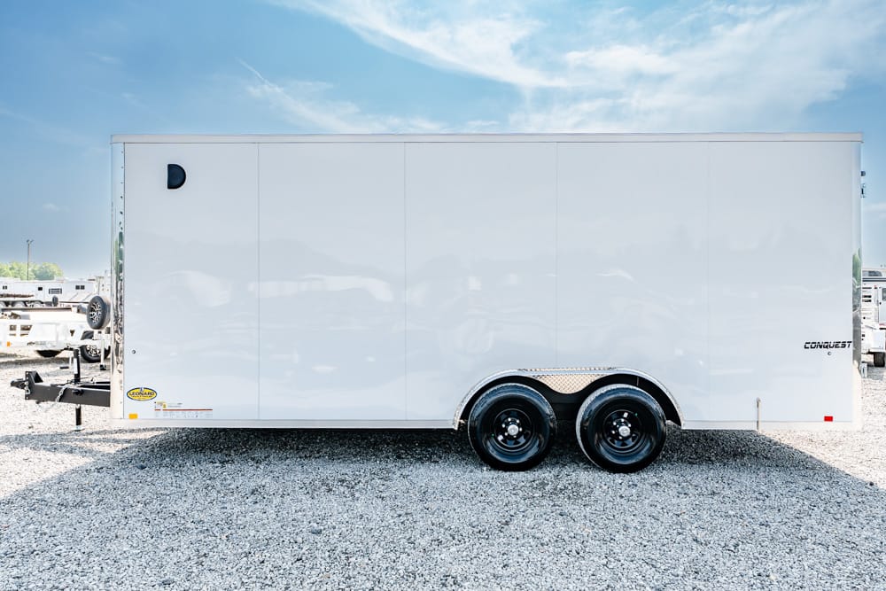White enclosed cargo trailer with dual rear wheels parked on a gravel lot beneath a blue sky