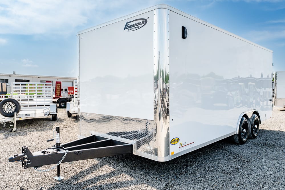 White enclosed cargo trailer with dual axles and a black hitch parked on a gravel lot, showing a front frame and chain.
