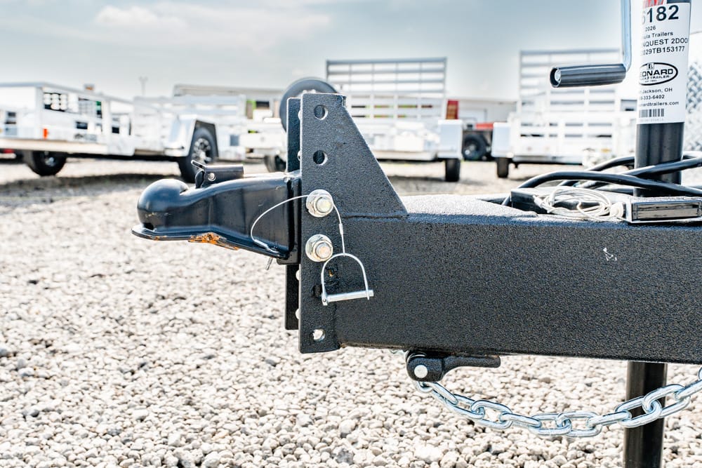 Close-up of a black trailer hitch and safety chain on a rugged trailer frame, set on gravel ground in a trailer yard.