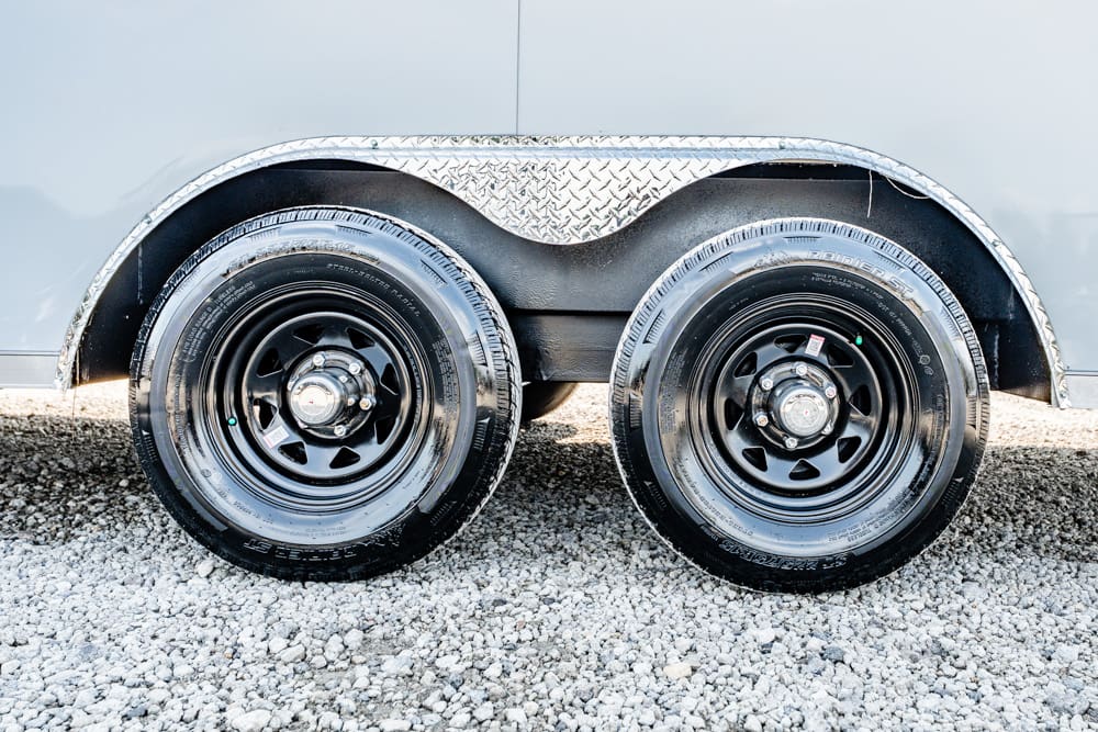 Twin trailer wheels with black rims and a diamond-plate fender, parked on gravel.