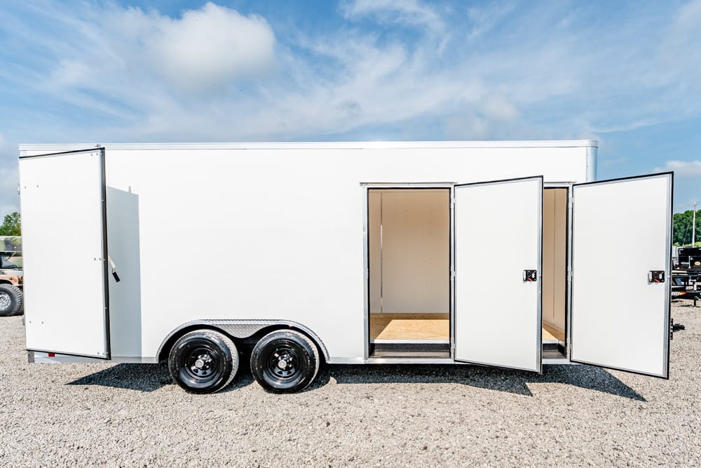 White cargo trailer on a gravel lot with three doors open, showing an empty wood-floored interior and dual axle wheels beneath a blue sky.