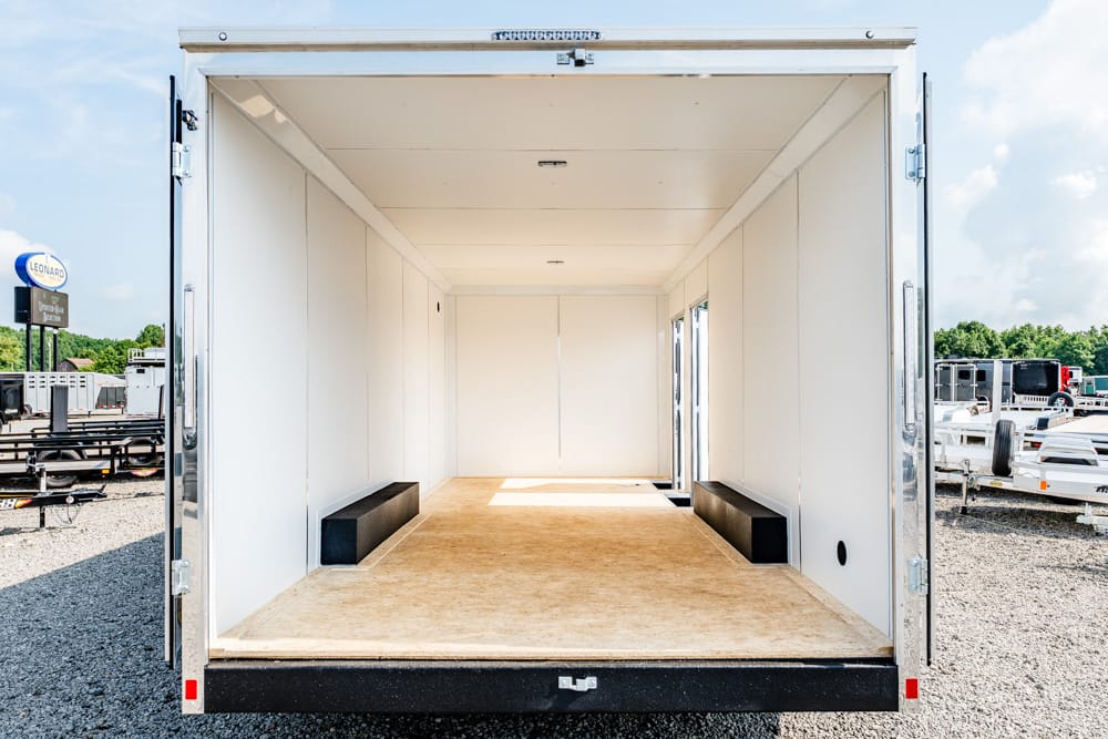 Empty white cargo trailer interior with a wooden floor and two black benches along both sides, doors open in a gravel lot.